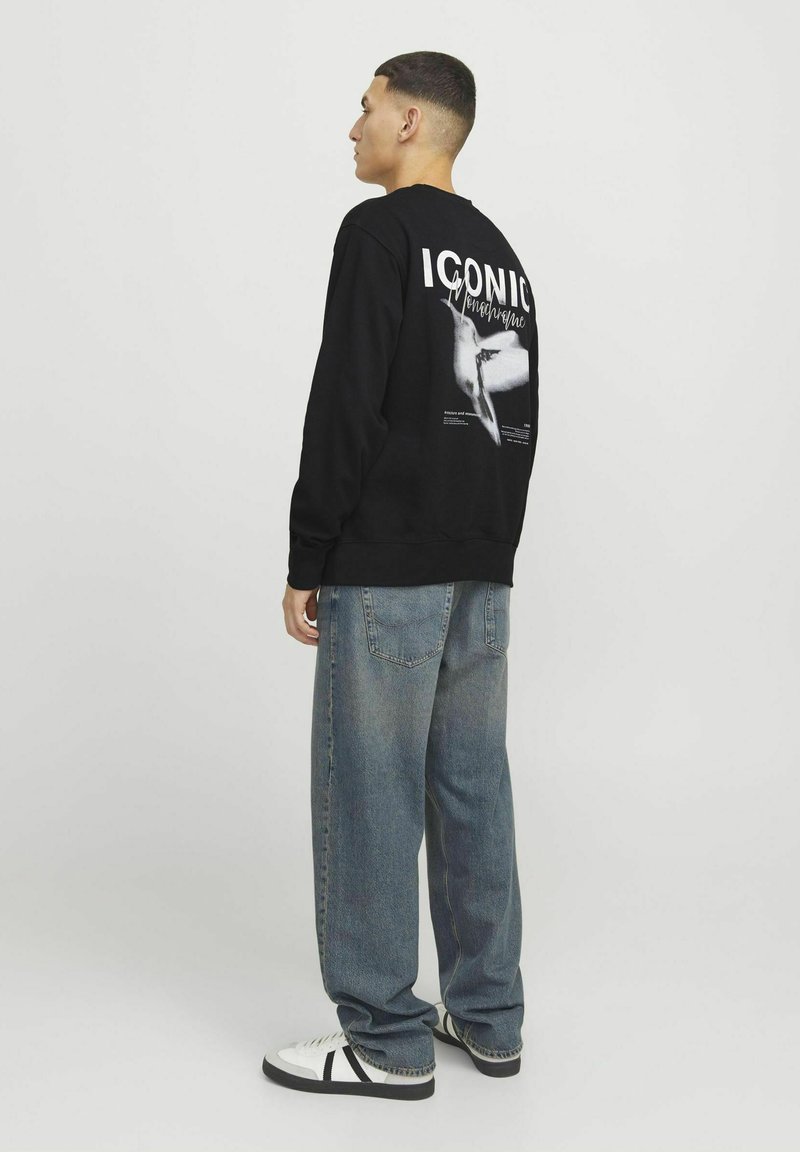 Sudadera negra con el texto "ICÓNICO" y un gráfico impreso en la parte de atrás. Combinada con unos pantalones de mezclilla sueltos de color azul claro y zapatillas blancas.