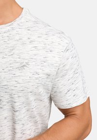 T-shirt gris clair à manches courtes en tissu texturé avec un motif subtilement moucheté. Présente un petit logo sur la poitrine.