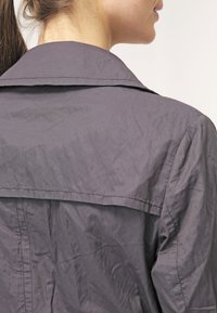 Femme à la peau claire et aux cheveux bruns attachés, portant une veste grise, vue de dos et de côté sur un fond uni.