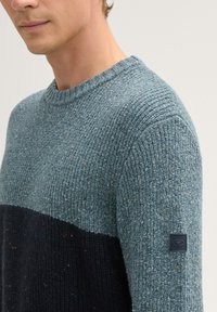 Strickpullover in Hellblau und Dunkelblau mit strukturierter Oberfläche. Verfügt über einen runden Ausschnitt und ein kleines Logo-Patch am linken Ärmel.