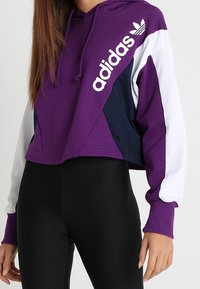 Hoodie accorciato con un corpo viola, pannelli blu navy e bianchi, cappuccio con cordino e logo "adidas" bianco sulla parte anteriore. Tessuto misto cotone.