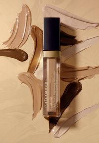Correcteur Futurist d'Estée Lauder dans un tube transparent avec un accent doré et un capuchon bleu marine, entouré de dégradés lisses de différentes teintes de fond de teint.