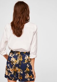 Nainen, jolla on pitkät ruskeat hiukset, pukeutuneena valkoiseen pitkähiharaiseen paitaan ja tummansinihin shortseihin, joissa on keltaisia ja vaaleanbeigejä kukkakuoseja, seisoo selkä kameraan päin.