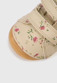 Brūnganas krāsas ādas kurpes ar ziedu rakstu rozā un zaļā krāsā, ar divām līplentas siksnām un teksturētu brūnganas krāsas zolīti. Tuvplāna skats.