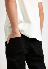 Černé džíny z denimu s přiléhavým střihem, se zadními kapsami a bílým bavlněným tričkem, viditelným na lemu. Textura vypadá hladce.