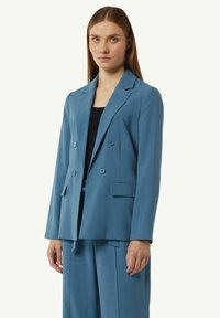 Błękitny dwurzędowy żakiet z wykładanym kołnierzem, dwiema przednimi kieszeniami i guzikami, wykonany z gładkiego materiału; zestawiony z czarną bluzką.