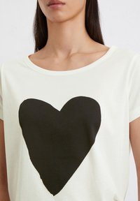 Witte katoenen t-shirt met een grote zwarte hartafbeelding in het midden, met een ronde halslijn en korte mouwen. Zachte textuur.