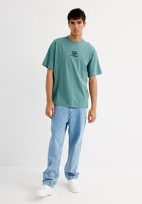 Camiseta de algodón verde menta con cuello redondo, mangas cortas y estampado de texto negro. Combinada con unos vaqueros de mezclilla claros y holgados y zapatillas blancas.