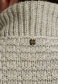 Maglione lavorato a maglia grigio chiaro con un motivo a diamante testurizzato, colletto alto e un'etichetta in metallo nero sul retro.