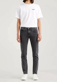 Jeans de mezclilla negros con ajuste slim, que cuentan con cinco bolsillos y cierre de botón. Combinados con una camiseta blanca lisa que muestra un pequeño logo.