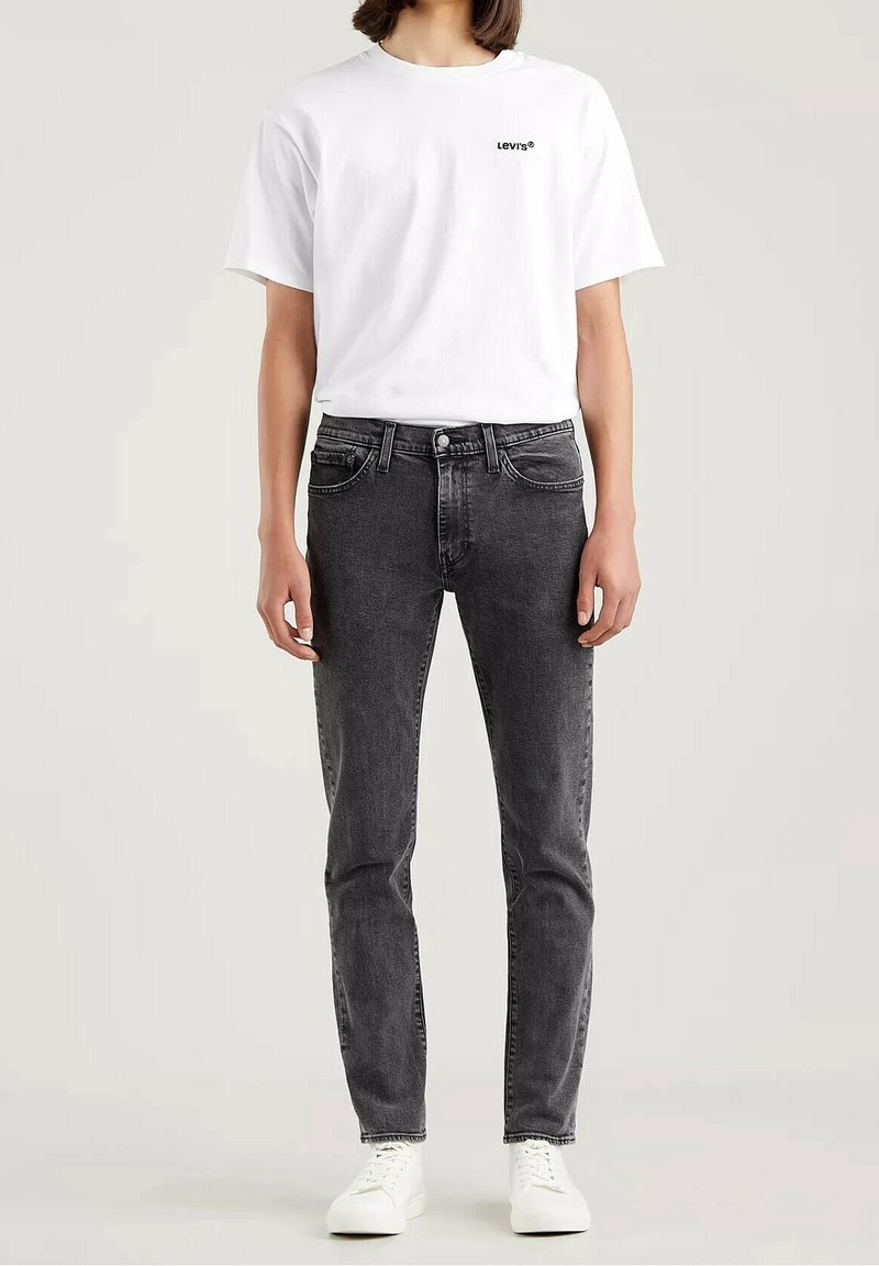Jeans de mezclilla negros con ajuste slim, que cuentan con cinco bolsillos y cierre de botón. Combinados con una camiseta blanca lisa que muestra un pequeño logo.