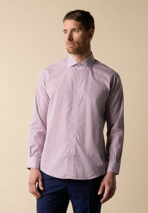 Mann, der ein langärmeliges, geknöpftes Hemd mit dünnen roten und weißen vertikalen Streifen und marineblaue Hose trägt, stehend vor einem beigen Hintergrund.