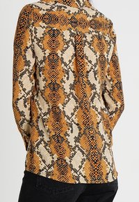 Camicia a maniche lunghe con motivo a stampa di serpente in arancione, nero e crema. Presenta una parte posteriore con bottoni e un tessuto testurizzato.