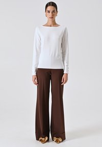 Maglietta bianca a maniche lunghe abbinata a pantaloni larghi marroni. La maglietta presenta un collo a barco e dettagli di bottoni discreti sulle maniche. Scarpe con accenti dorati.