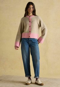 Žena stojaca v béžovom cardigáne s ružovým lemom a gombíkmi, s manžetovými modrými džínsami, vzorovanými ponožkami a bielymi teniskami na jednoduchom pozadí.
