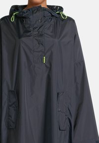 Poncho de pluie navy fabriqué en matériau imperméable, avec une capuche, des fermetures à pression et deux poches latérales avec rabats et boutons. Accents néon inclus.