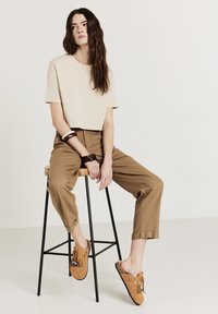 T-shirt beige corto, pantaloni a pieghe color beige, e scarpe slip-on in suede marrone con nappe e perline decorative. Seduto su uno sgabello di legno.