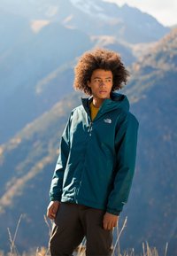 Veste imperméable teal avec capuche, poignets élastiques et poches latérales, associée à une chemise jaune et un pantalon sombre dans un cadre extérieur montagneux.