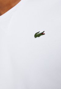 Vit bomull T-shirt med V-ringning och en liten, grön broderad krokodil-logotyp placerad på vänster sida av bröstet.