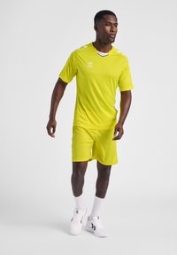 Neongele sporttrui met bijpassende shorts, ademend materiaal, korte mouwen, V-hals en contrasterende witte accenten. Draagster draagt witte sneakers.