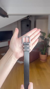 Fekete bőr öv kézben tartva, ezüst színű dupla csattal és három fémes hurkokkal. Sima felületű, minimalista dizájn.