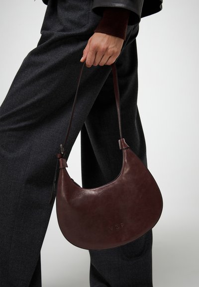 Borsa in pelle bordeaux con design curvo, tracolla regolabile e logo impresso, tenuta da una persona che indossa pantaloni grigi.