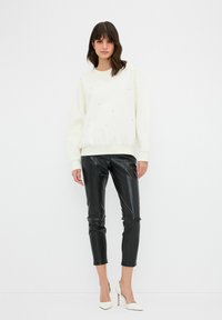 Bílá mikina se strukturálními ozdobami, spárovaná s černými kalhotami z umělé kůže, doplněná bílými lodičkami na vysokém podpatku.