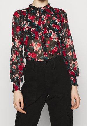 Przezroczysta czarna bluzka z czerwonym i różowym kwiatowym wzorem, zawiązywany kołnierzyk, bufiaste rękawy, zestawiona z czarnymi, wysokim stanem spodniami z kieszeniami cargo.