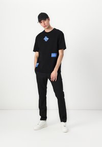 Svart T-skjorte med korte ermer og blå grafiske lapper, kombinert med svarte bukser og hvite joggesko. Antrekket har et uformelt, moderne design.