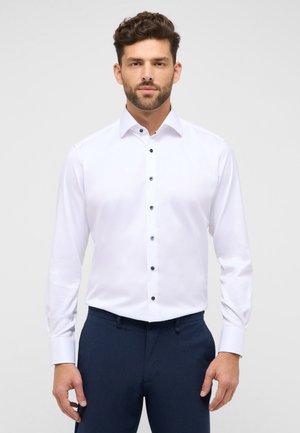 Witte lange mouwen shirt met een vaste textuur, met een puntige kraag en zes donkerblauwe knopen aan de voorkant. Gecombineerd met donkere broeken.