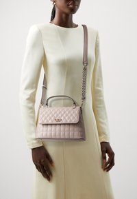 Přebalovaná kabelka ve světlém taupe se strukturovaným tvarem, horním úchytem a odnímatelným řetízkovým popruhem. Má klopnové zapínání a kovový detail loga.