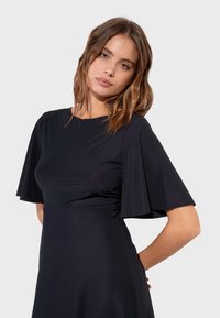 Svart knälång klänning med fladdrande ärmar, mjukt tyg, rund halsringning och figursydd midja. Designen är enkel och elegant.