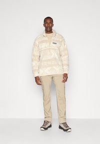 Krémový fleece pulovr s geometrickým vzorem, límcem na půl zip a logem. V kombinaci s béžovými kalhotami a botami s tmavými akcenty.