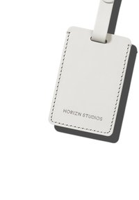 Weißes Ledergepäckanhänger mit gesticktem Rand. Verfügt über eine oberseitige Schlaufe zur Befestigung und dem Aufdruck "HORIZN STUDIOS" in Grau.