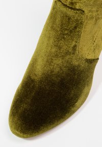 Chaussure en velours vert olive avec un bout arrondi et des coutures visibles sur le dessus, photographiée sur fond blanc.