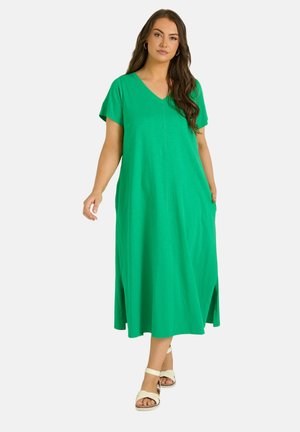 Frau steht in einem leuchtend grünen, kurzärmeligen Midi-Kleid mit V-Ausschnitt und seitlichen Schlitzen, trägt weiße offene Sandalen mit Knöchelriemen.