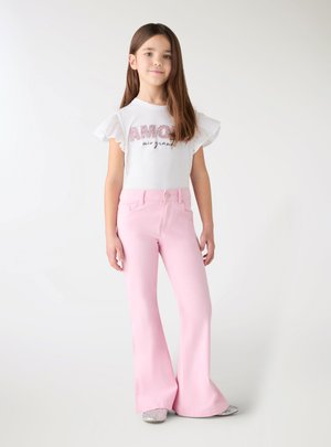 Dziewczyna stoi, ubrana w białą koszulę z falbankami na rękawach z napisem "Amour", jasnoróżowe rozszerzane spodnie i błyszczące srebrne buty na białym tle.