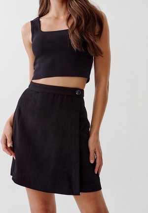 Femme portant un crop top noir sans manches et une jupe noire taille haute avec un détail de bouton sur le côté, sur un fond uni.
