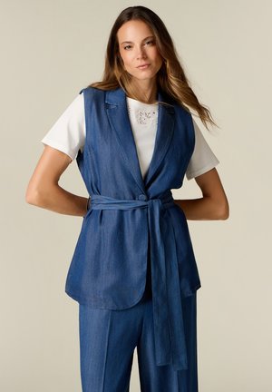 Vrouw draagt een blauwe denim mouwloze gilet met riem over een wit kort mouw topje met bijpassende blauwe denim broek, staand met handen in de zij.