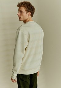 Felpa color beige chiaro con texture, maniche lunghe e collo tondo, abbinata a pantaloni verde scuro, vista da dietro su uno sfondo neutro.