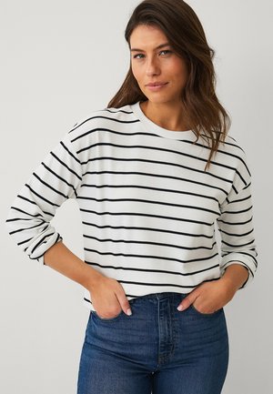 Kvinde med langt brunt hår iført en hvid langærmet skjorte med sorte vandrette striber og blå højtaljede jeans, hænderne i lommerne.