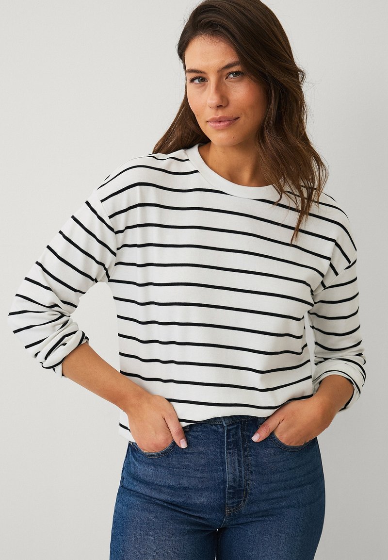 Femme aux longs cheveux bruns portant une chemise blanche à manches longues avec des rayures horizontales noires et un jean bleu taille haute, les mains dans les poches.