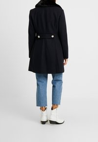 Cappotto in misto lana blu navy con colletto in pelliccia sintetica, dettaglio in vita sblusato e bottoni argentati, abbinato a jeans cropped azzurri chiari e stivali bianchi.