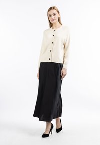 Cardigan in maglia beige chiaro con scollo rotondo e bottoni neri, abbinato a una lunga gonna nera in satin e tacchi a spillo neri.