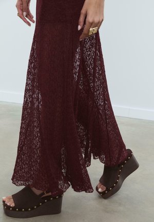 Burgundy lace dress flows to the floor, paired with dark wedge sandals featuring gold stud accents and a smooth wooden platform.