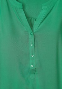 Grön blus med v-ringning, som har en rad knappar fram, mjukt, genomskinligt tyg och subtil textur.