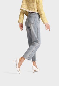 Jeans azzurri a vita alta con una vestibilità rilassata, orlo strappato, abbinati a tacchi bianchi in pelle lucida con accenti dorati.