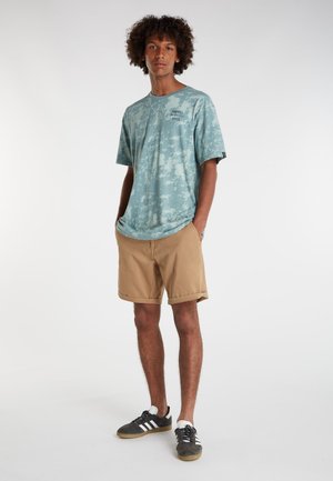 Hellblaues Batik-T-Shirt mit kurzen Ärmeln, kombiniert mit khakifarbigen Shorts. Das Model trägt schwarze Sneakers mit weißen und grauen Akzenten.