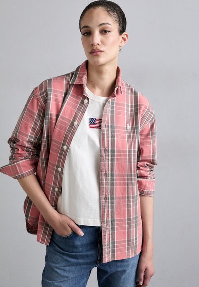 Camisa de cuadros rosa y gris con botones sobre una camiseta blanca con una pequeña bandera americana bordada, combinada con jeans de denim azul.