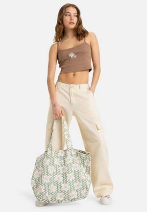 Grande borsa tote con un motivo floreale verde e bianco, dotata di manici robusti e una etichetta con logo. Indossata con un top corto marrone e pantaloni chiari.
